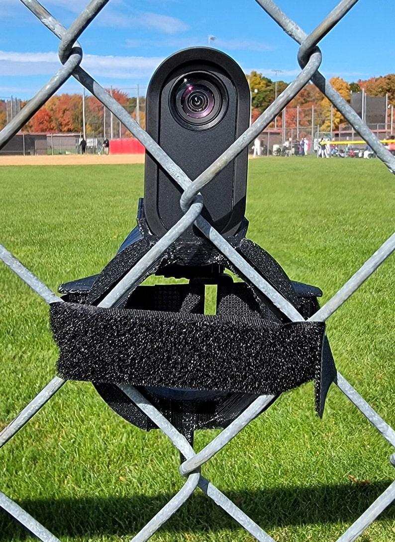 May include: A black camera mounted on a chain link fence with a black strap. The camera is pointed towards a baseball field.
