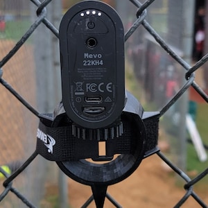 May include: A black Mevo 22KH4 camera with a black mounting bracket attached to a chain link fence. The camera has a power button and a USB-C port. The camera is turned on and the lights are on.