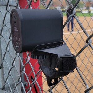 May include: A black camera with a black strap attached to a metal fence. The camera has a black and white label that says "Made in China" and "FCC CE".