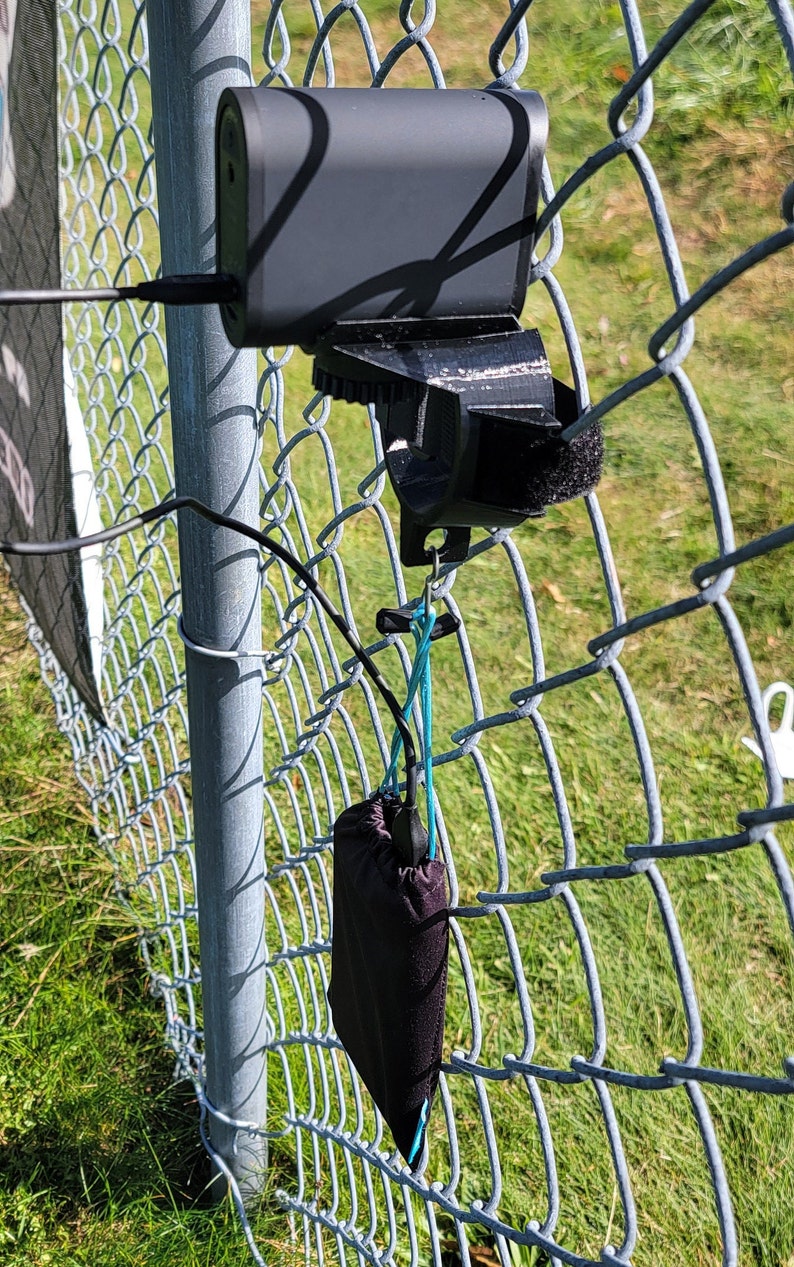 May include: A black power bank attached to a chain link fence with a black pouch hanging below it.