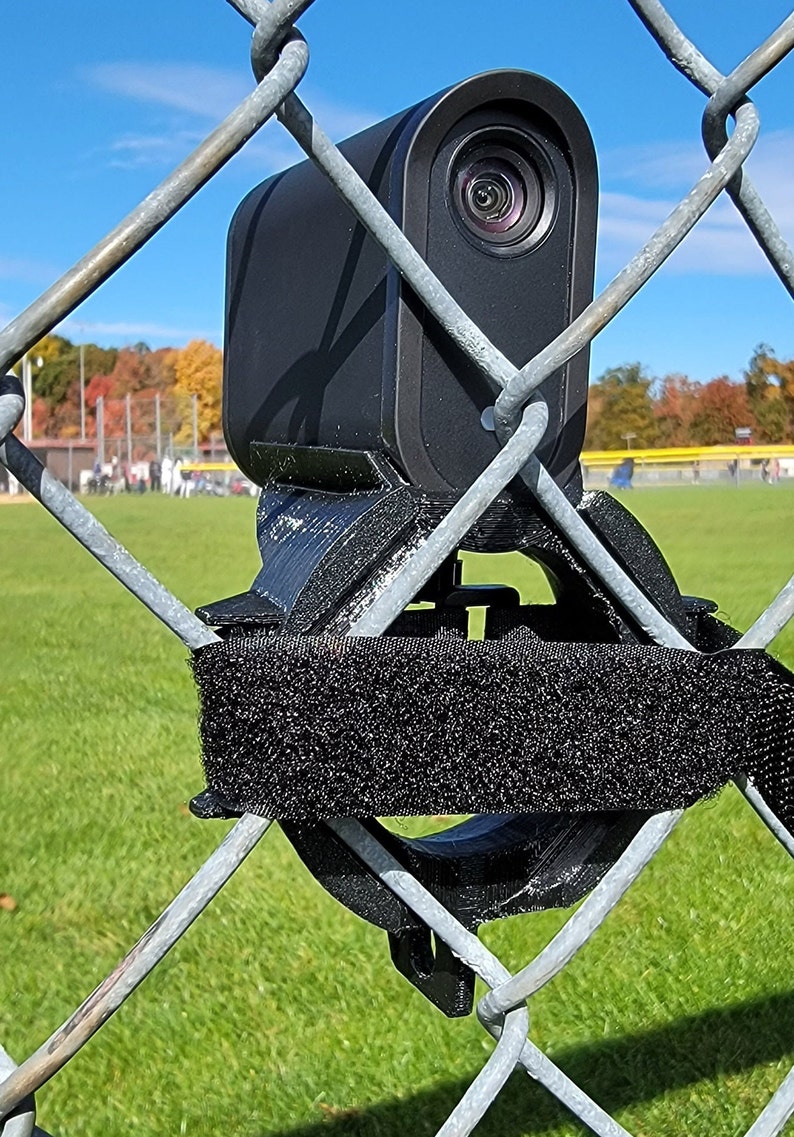 May include: A black camera mounted on a chain-link fence with a black strap securing it. The camera is pointed towards a baseball field.