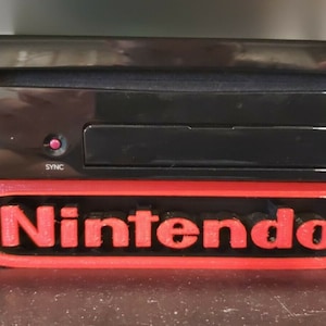 Puede incluir: Una consola Nintendo Wii U negra con una luz roja en la parte delantera, sentada en un soporte rojo con la palabra "Nintendo" impresa en él.