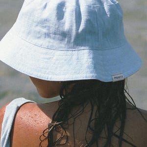 May include: A person wearing a light blue linen bucket hat with the word "maarts" on the side. The person's hair is wet and dark, and their shoulders are visible.