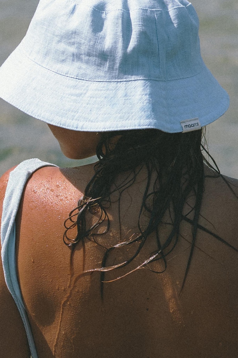 May include: A person wearing a light blue linen bucket hat with the word "maarts" on the side. The person's hair is wet and dark, and their shoulders are visible.