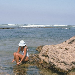 May include: A woman wearing a white swimsuit and a light blue hat sits on a rock by the ocean. The water is clear and blue, and the sky is a light blue.