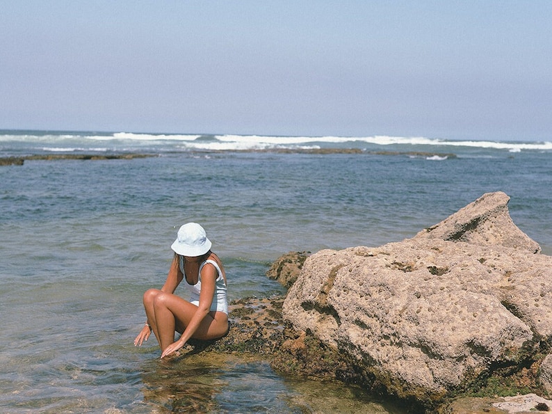 May include: A woman wearing a white swimsuit and a light blue hat sits on a rock by the ocean. The water is clear and blue, and the sky is a light blue.
