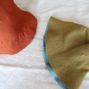 Puede incluir: Dos sombreros de cubo de lino. Uno es de color naranja &oacute;xido s&oacute;lido. El otro es amarillo mostaza con un ribete azul turquesa.
