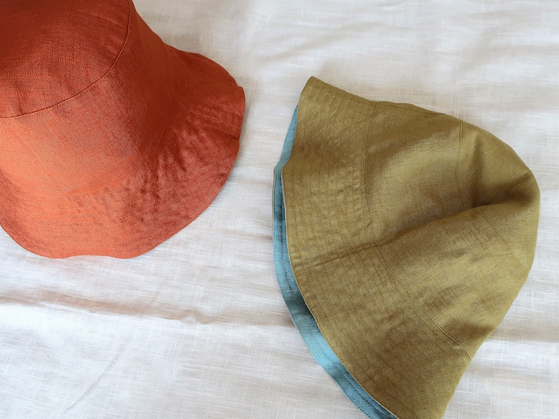 Puede incluir: Dos sombreros de cubo de lino. Uno es de color naranja &oacute;xido s&oacute;lido. El otro es amarillo mostaza con un ribete azul turquesa.