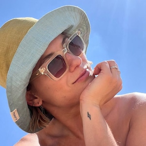 Op de afbeelding: Een persoon draagt een lichtblauwe en gele bucket hat met een transparante zonnebril. De hoed heeft een klein label met de tekst "rokit" aan de zijkant. De persoon heeft een bliksemschicht tatoeage op de pols.