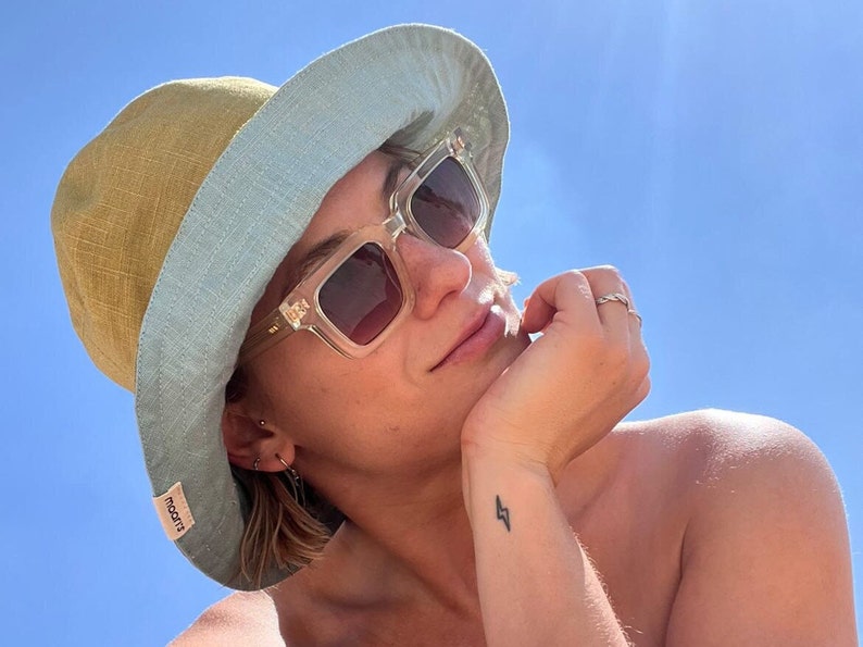 Op de afbeelding: Een persoon draagt een lichtblauwe en gele bucket hat met een transparante zonnebril. De hoed heeft een klein label met de tekst "rokit" aan de zijkant. De persoon heeft een bliksemschicht tatoeage op de pols.