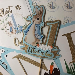 Peut inclure: Une bannière de fête bleu clair et dorée avec le texte "Elliot is One". Elle présente une illustration de Pierre Lapin et le chiffre 1. Une carotte et une clôture blanche sont également visibles.