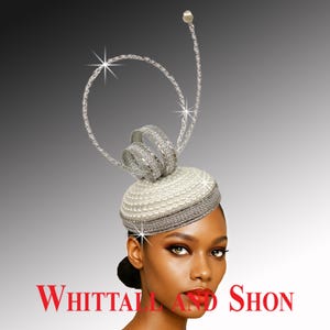 May include: A white pillbox hat adorned with rows of small white pearls and a silver metallic band. The hat features a tall, circular silver wire structure with a textured finish and a single pearl at the top. The text WHITTALL AND SHON is visible.