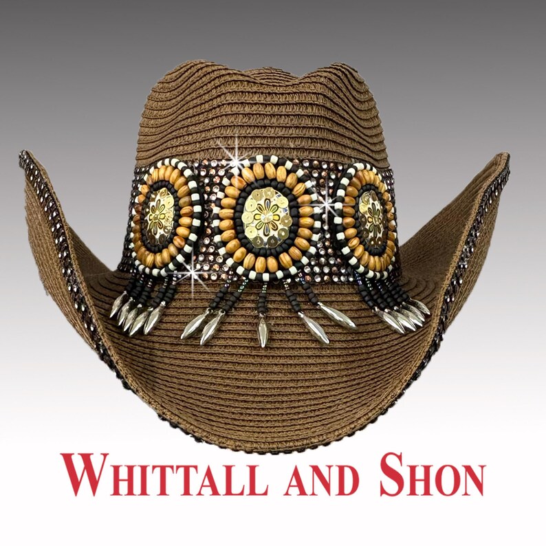 May include: Brown cowboy hat with decorative embellishments. The hat features a band adorned with gold, black, and white beads, sequins, and dangling silver-coloured charms. The brim is trimmed with small, dark beads. The text "WHITTALL AND SHON" is displayed in red.