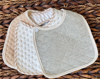 Può includere: Un bavaglino per neonato beige e grigio con un design trapuntato e una chiusura a scatto. Il bavaglino è realizzato in tessuto morbido e assorbente, perfetto per proteggere i vestiti del bambino da schizzi e sbavature.