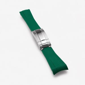 Puede incluir: Una correa de reloj de goma verde con un cierre plateado. La correa es curva y se estrecha hacia los extremos. El cierre es rectangular y tiene un acabado pulido.
