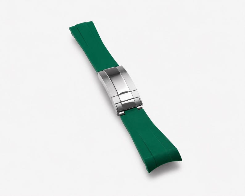 Puede incluir: Una correa de reloj de goma verde con un cierre plateado. La correa es curva y se estrecha hacia los extremos. El cierre es rectangular y tiene un acabado pulido.