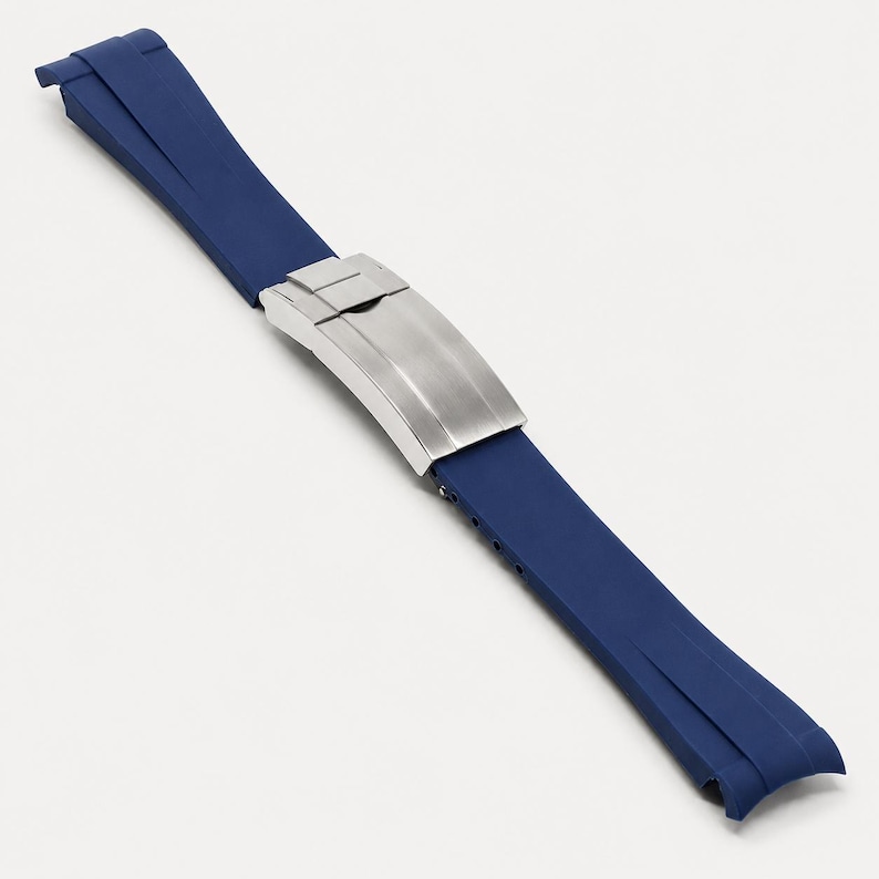 Puede incluir: Una correa de reloj de goma azul marino con un cierre de acero inoxidable. La correa es de color azul oscuro y tiene una textura suave. El cierre es de color plateado cepillado y tiene forma rectangular.