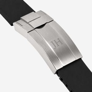 Puede incluir: Primer plano de un cierre de reloj con las iniciales "JH" grabadas en la superficie de metal cepillado. El cierre est&aacute; conectado a una correa de reloj de goma negra, mostrando un dise&ntilde;o moderno.