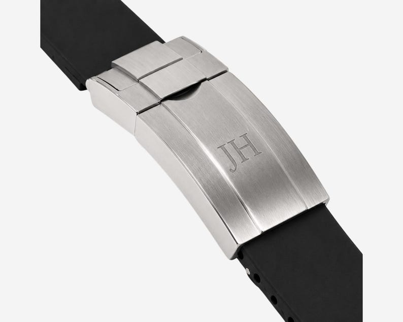 Puede incluir: Primer plano de un cierre de reloj con las iniciales "JH" grabadas en la superficie de metal cepillado. El cierre est&aacute; conectado a una correa de reloj de goma negra, mostrando un dise&ntilde;o moderno.