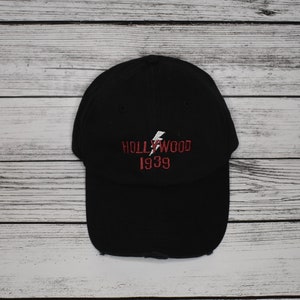 Puede incluir: Gorra de béisbol negra con un rayo blanco y texto bordado en rojo que dice "HOLLYWOOD 1939".