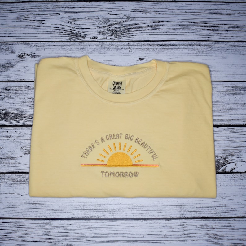 Puede incluir: Una camiseta amarilla con un amanecer bordado y el texto "There's a great big beautiful tomorrow".