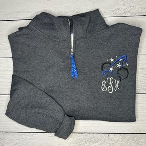 May include: A dark gray fleece pullover with a half-zip closure. The pullover features an embroidered design of a Mickey Mouse head wearing a sorcerer's hat with stars. The initials "E.F.K." are embroidered below the Mickey Mouse design.