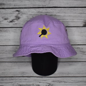 May include: A purple bucket hat with a yellow embroidered sun design featuring a frying pan inside. The hat is on a black hat stand.