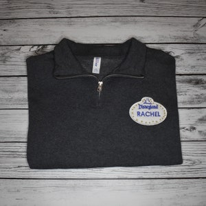 May include: A dark gray 1/4 zip sweatshirt with a white and gold Disneyland name tag patch that says "Disneyland" and "RACHEL" in blue text.