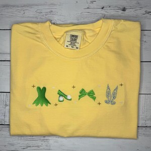 Puede incluir: Camiseta amarilla Comfort Colors con diseños bordados de vestido verde, zapato, lazo y alas de hada. La camiseta tiene cuello redondo y una etiqueta que dice "Comfort Colors" y "XL - TG - EG".