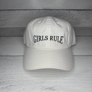 May include: White baseball cap with the text "GIRLS RULE" embroidered in black. The cap has a curved brim and an adjustable strap at the back. The hat is made of a soft, durable material.