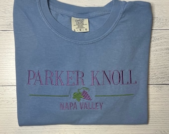 Puede incluir: Camiseta azul claro con bordado morado "PARKER KNOLL", un motivo de uvas y "NAPA VALLEY". La camiseta está doblada, con una etiqueta "Comfort Colors". El diseño probablemente está relacionado con el vino.