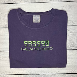 May include: Purple t-shirt with green embroidered text that reads "999999 GALACTIC HERO".