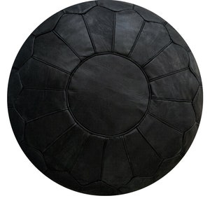 Op de afbeelding: Een ronde, zwarte leren poef met een gestikt, gesegmenteerd ontwerp. De poef heeft een centraal cirkelvormig paneel en uitstralende bloembladachtige secties, waardoor een decoratief en functioneel meubelstuk ontstaat. De donkere kleur en textuur suggereren duurzaamheid en stijl.