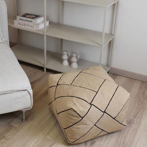 Op de afbeelding: Een vierkante vloerpoef met een gestructureerd beige stoffen oppervlak en een opvallend zwart gestikt ruitpatroon. De poef heeft een zachte, licht afgeronde vorm en is gemaakt van geweven materiaal met een geometrisch ontwerp.