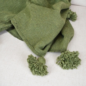 May include: A green knitted throw blanket with fringe tassels. The blanket is folded and draped over a light-coloured surface.