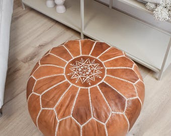 May include: A round, brown leather pouf with white stitching and a central starburst design. The pouf is a decorative piece of furniture, likely used as a footrest or extra seating. The design is geometric and the color is a warm, earthy tone.