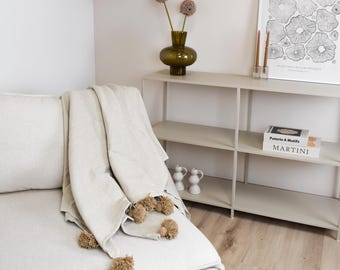 Puede incluir: Una manta de color beige claro con pompones decorativos de color tostado a lo largo del borde, colocada sobre un sofá de color claro. La manta tiene una textura de tela y se encuentra en una habitación con una estantería de metal y un cuadro.
