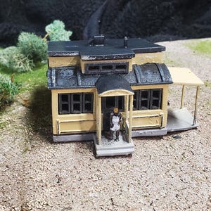 Puede incluir: Modelo en miniatura de una casa amarilla con tejado y molduras negras. La casa tiene varias ventanas y un pequeño porche. Una figura en miniatura está de pie en la puerta. El modelo está sobre una superficie texturizada.