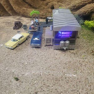 Könnte beinhalten: Miniatur-Diorama-Szene mit einem Diner mit grauem Dach und leuchtendem violettem Innenraum. Zwei Oldtimer, einer gelb und einer blau, sind in der Nähe geparkt. Kleine Figuren sitzen an Picknicktischen.