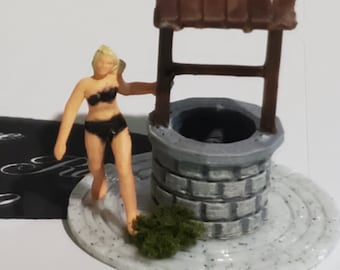 May include: A miniature scene featuring a gray stone well with a brown wooden roof. A small figure in a black bikini stands near the well. The well sits on a white, speckled base with a patch of green foliage.