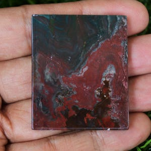 May include: A square, red, green and black agate cabochon with a swirling pattern. The cabochon is polished and has a smooth surface.