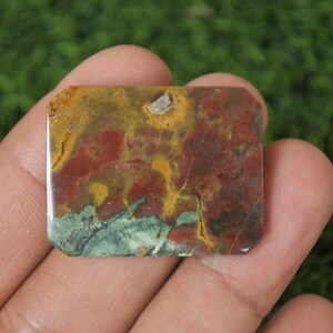 May include: A rectangular gemstone with a mix of colors, including red, yellow, and green. The stone has a unique pattern and is held in a hand. The gemstone is likely used for jewelry or decorative purposes.