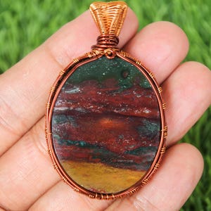 May include: Oval pendant with a copper wire wrap. The stone features a mix of colors, including red, green, and yellow. The pendant is held in a hand against a green background.