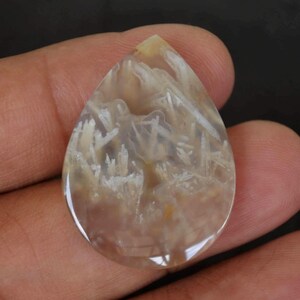 Könnte beinhalten: Ein tropfenförmiger Achat-Cabochon mit einem weißen und grauen dendritischen Muster. Der Achat ist durchscheinend und hat eine glatte, polierte Oberfläche.