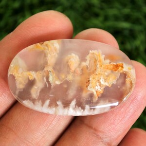 Könnte beinhalten: Ovale Edelstein mit einer durchscheinenden, hellrosa Basis. Der Stein weist komplizierte Muster aus Orange, Weiß und Braun auf, die einer Landschaft oder Wolkenformation ähneln. Die polierte Oberfläche reflektiert das Licht und hebt die natürlichen Variationen hervor.