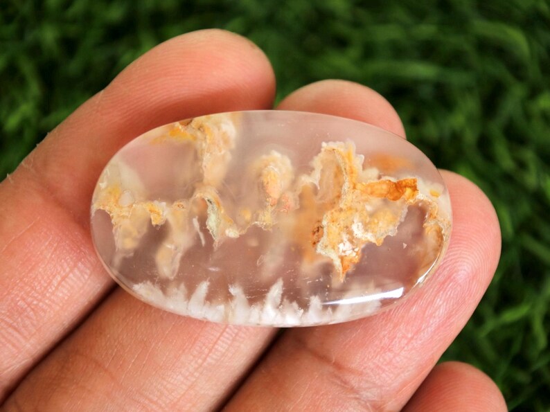 May include: An oval gemstone with a translucent, pale pink hue. It displays detailed patterns in shades of orange, white, and brown, reminiscent of a landscape. The polished surface reflects light, showcasing the natural variations in the stone.