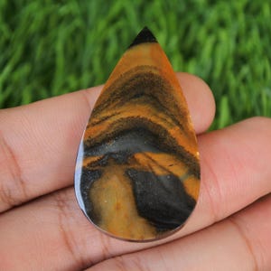 May include: A teardrop-shaped cabochon gemstone with a swirling pattern of black, brown, and yellow.