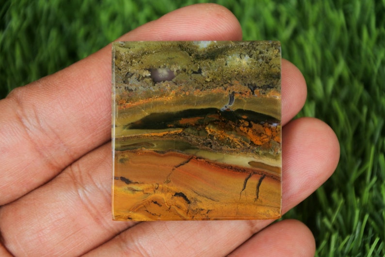 May include: A square-shaped, polished, multi-coloured agate stone with brown, yellow, green, and black banding. The stone has a natural, organic pattern.