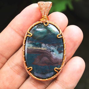 May include: A copper wire wrapped pendant featuring a green, blue, and purple agate stone. The stone has a swirling pattern and is set in a copper wire frame.