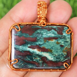 Peut inclure: Pendentif rectangulaire avec une monture et une bélière en fil de cuivre. La pierre à l'intérieur présente un motif moucheté de couleurs sarcelle, marron et crème. Le pendentif est tenu dans une main sur un fond vert flou.